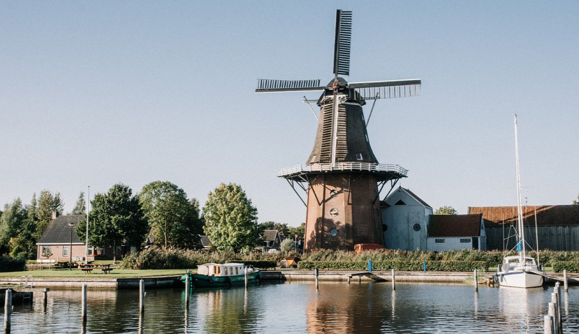 Traditional windmill by the water with boats moored nearby, under a blue sky in a serene setting.