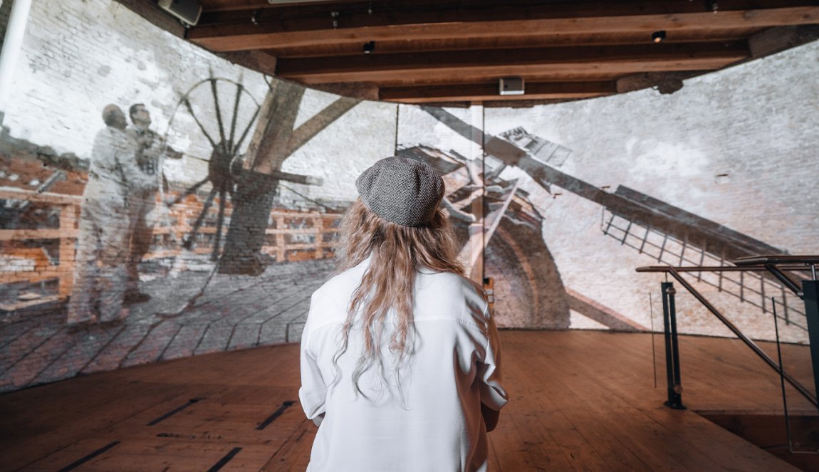 A person in a white shirt and beret looks at a historical photo projection of two men on top of a windmill.