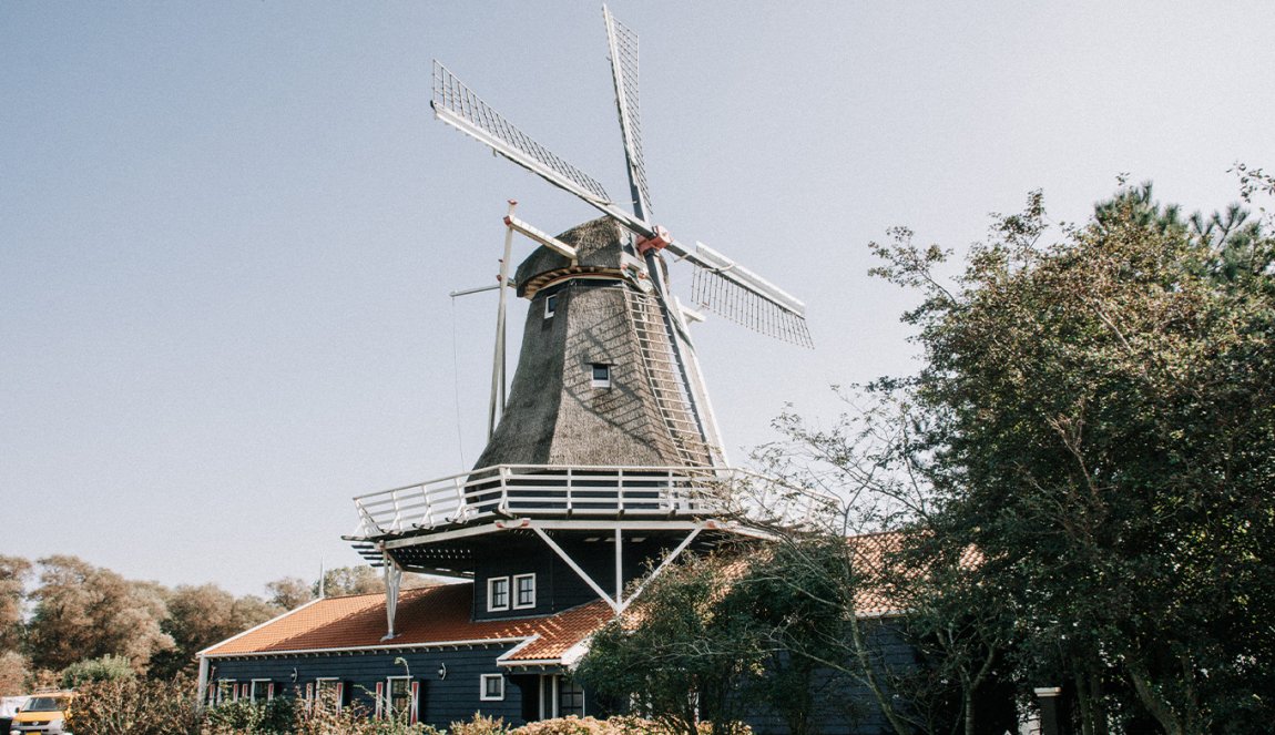 A classic Dutch windmill with a thatched roof and white sails, set against a clear sky, surrounded by greenery.