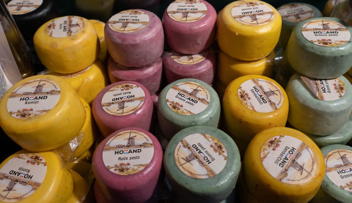 Colorful mini cheese wheels labeled with varieties like 'Komijn' and 'Groene pesto', displayed in a cheese shop.