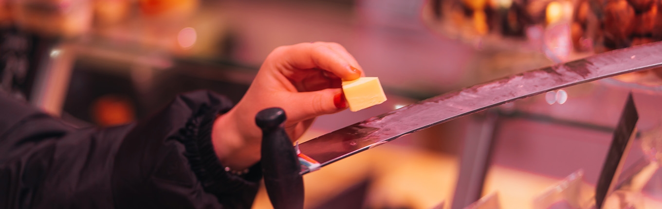 A hand holding a piece of cheese on a spatula at a tasting market, with other cheeses softly blurred in the background.
