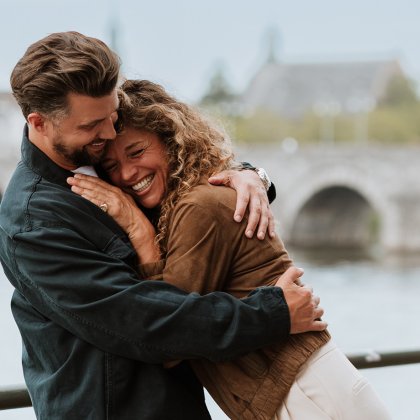 Couple embracing along the River Meuse in Maastricht, with the historic Sint Servaas Bridge in the background, capturing a romantic city break in the Netherlands