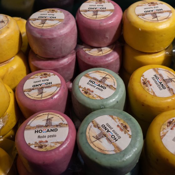 Colorful mini cheese wheels labeled with varieties like 'Komijn' and 'Groene pesto', displayed in a cheese shop.