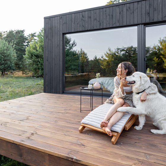 Woman sits on sunbed with dog on wooden terrace near the modern vacation house