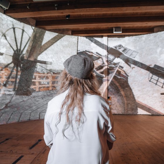 A person in a white shirt and beret looks at a historical photo projection of two men on top of a windmill.